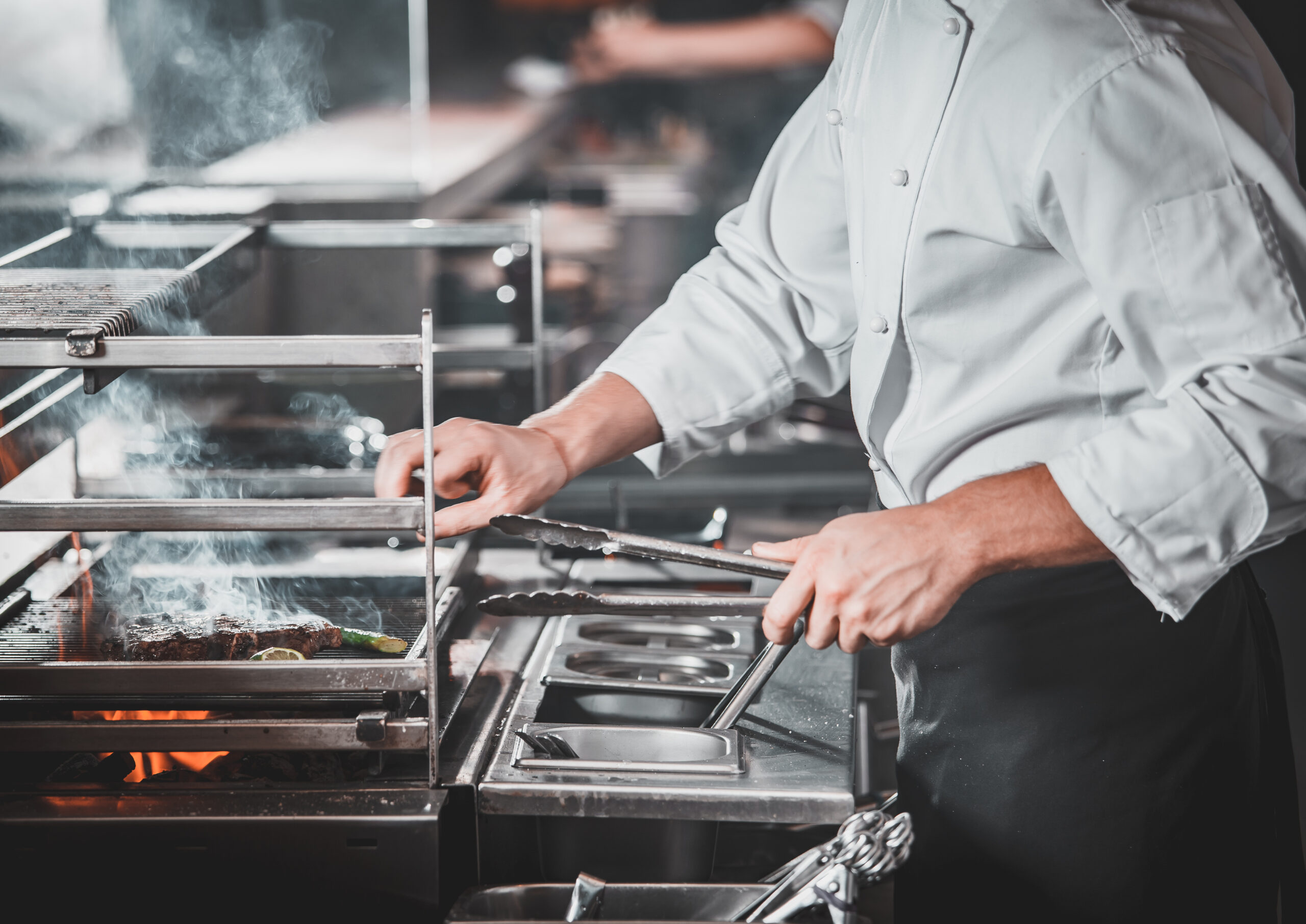 White Chef In Apron Standing Near The Brazier Whith Coals, Only Hands. Man Cooking Beef Steak In The Interior Of Modern Professional Kitchen